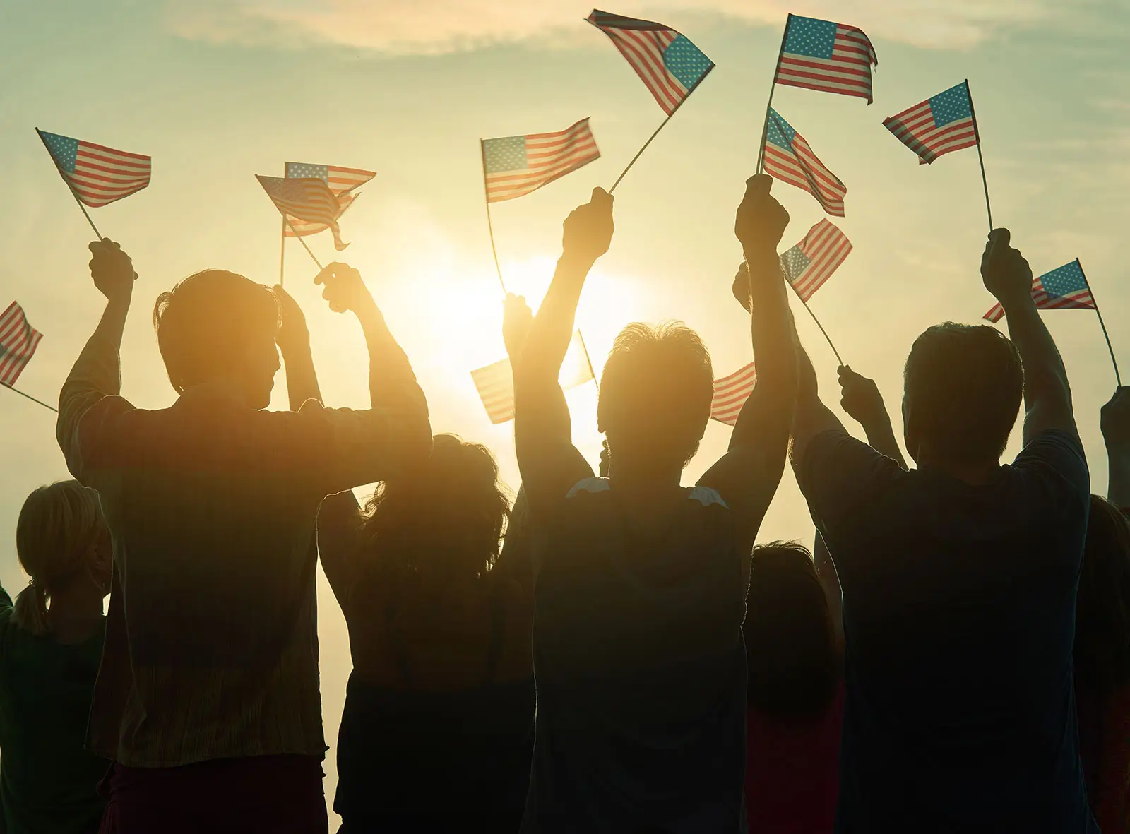 A group of people are seen in silhouette against a bright, setting sun, enthusiastically waving numerous American flags. Their arms are raised high, indicating celebration, patriotism, or a demonstration. The golden light of the sun creates a warm and uplifting atmosphere. The image evokes feelings of national pride and unity.